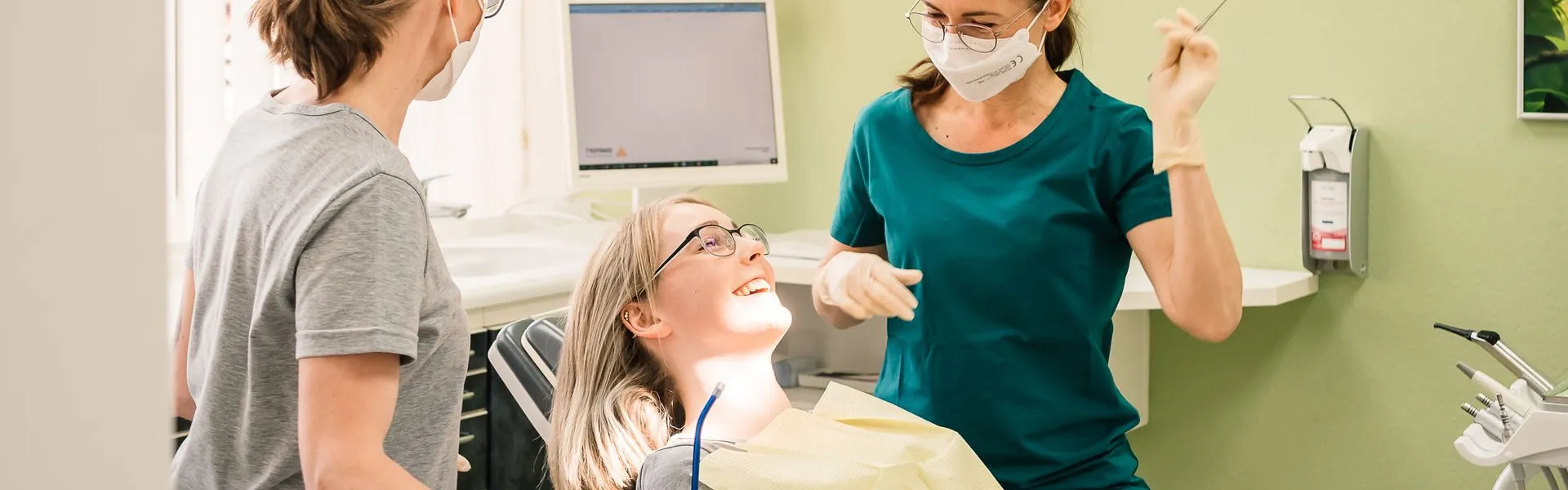 Eine lachende Patientin mit Brille liegt im Behandlungsstuhl, zwei Mitarbeiterinnen mit Mundschutz in grüner und grauer Kleidung sprechen freundlich mit ihr.
