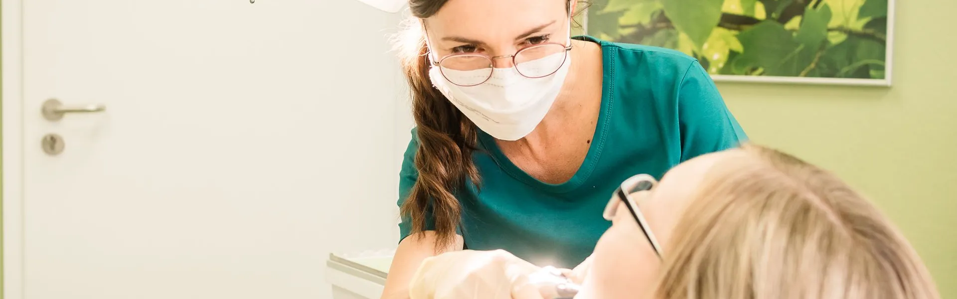 Zahnärztin mit Mundschutz und Brille behandelt konzentriert eine Patientin mit Brille in einem hellen Behandlungsraum mit grüner Wand.