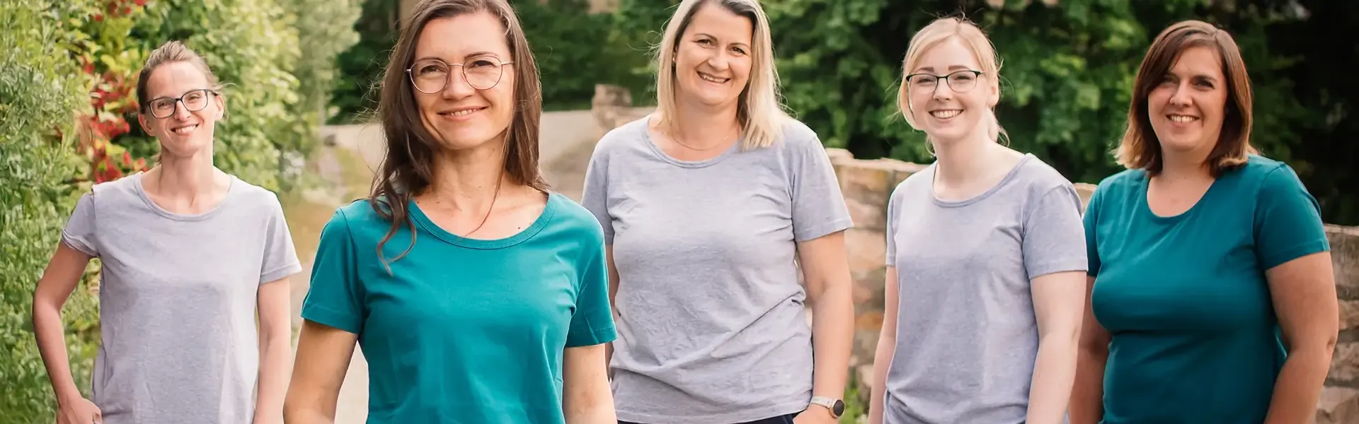 Fünf lächelnde Frauen in grauen und türkisfarbenen T-Shirts stehen nebeneinander im Freien vor Grün und einer Steinmauer.