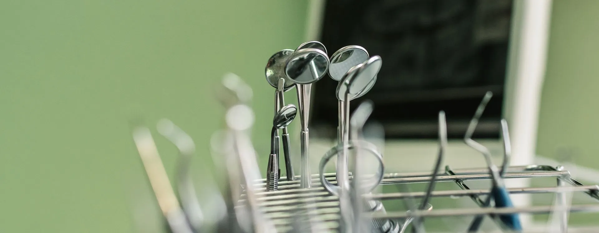 Zahnärztliche Mundspiegel und Instrumente stehen in einem Metallständer vor einer grünen Wand, der Hintergrund ist weich unscharf.
