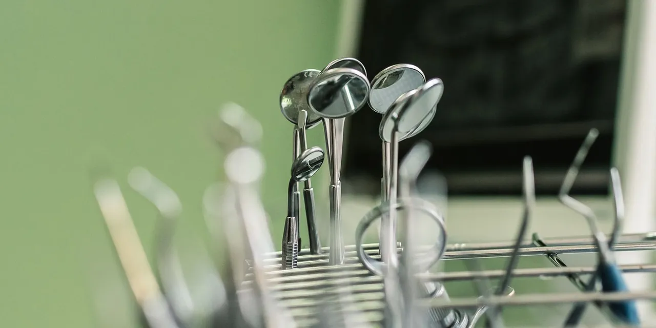 Zahnärztliche Mundspiegel und Instrumente stehen in einem Metallständer vor einer grünen Wand, der Hintergrund ist weich unscharf.