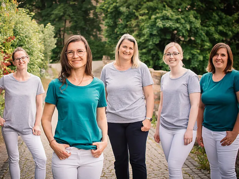 Fünf lächelnde Frauen in grauen und petrolfarbenen Shirts stehen im Freien auf einem Kopfsteinpflasterweg vor Grün und einer Steinmauer.